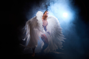 Handsome young athletic man with a bare torso who looks like an angel with white wings. Model dancer posing in a dark studio on black background