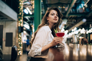 Portrait of trendy dressed female diva looking at camera during cocktail time in local bar restaurant, beautiful Caucasian woman with alcohol beverage posing during weekend leisure in public cafe