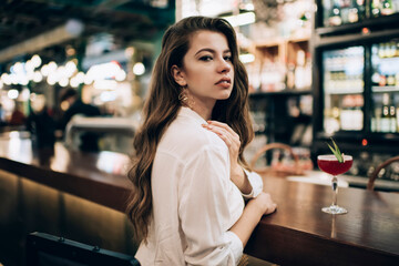 Portrait of trendy dressed diva woman looking at camera during cocktail time in local bar restaurant, attractive Caucasian customer with alcohol beverage posing during weekend leisure in public cafe