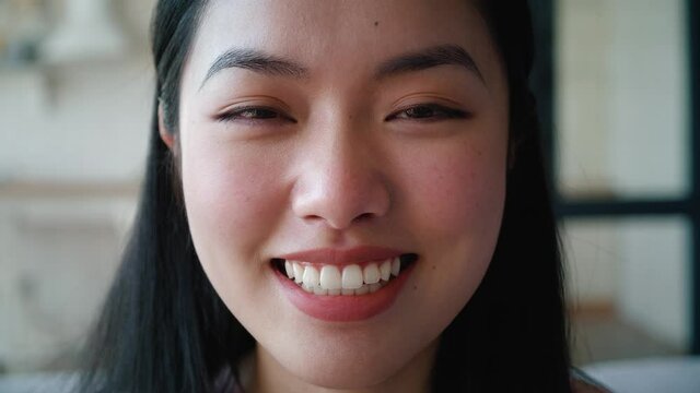 Close-up. Cropped confident face portrait of gorgeous young Asian woman smiling while posing to camera. Focus on the calm look of a serene woman with toothy smile, looking confidently into the camera