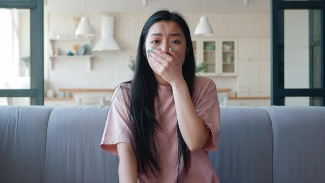 Young dark-haired with long hair woman of Asian descent looks at camera, sitting on sofa and suddenly covers her mouth being afraid and shocked by watched. Main focus on facial expression and emotion