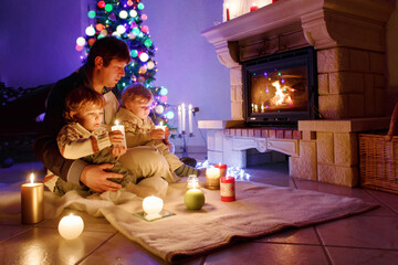 Father and two little toddler boys sitting by chimney, candles and fireplace and looking on fire.