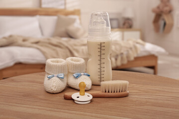 Fototapeta na wymiar Baby booties, pacifier, feeding bottle and brush on wooden table in bedroom. Maternity leave concept