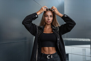 Fashion young pretty girl with a beautiful body in a fashionable denim jacket with rhinestones in black jeans and tank top posing in the city and looks at the camera