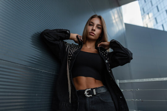 Fashion young model woman in a black stylish jeans jacket with rhinestones in a black t-shirt top posing near a dark metal wall