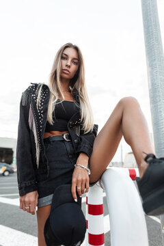 Young beautiful sexy woman with beautiful slim legs in a black denim jacket with stylish jeans shorts with a black cap and leather shoes posing in the city. Urban women's style look