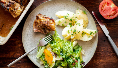 Boiled young potatoes with dill, fresh vegetable salad and baked chicken thighs on a dark wooden background.