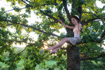 beautiful young girl in summer dress and hat is sitting on tree, on branch of large oak tree in sunset rays and smiling and enjoying evening nature in open air. Summer holidays in nature