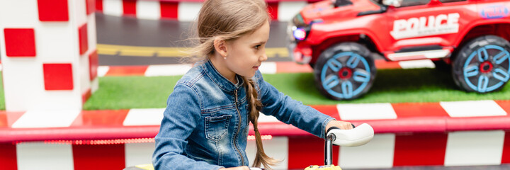 little happy kid girl fun riding an small electric cars on sport ground in a playground for entertainments. children riding in the toy auto in an amusement park. banner