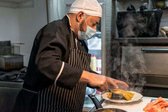 Ethnic chef putting delicious cannelloni on plate in kitchen
