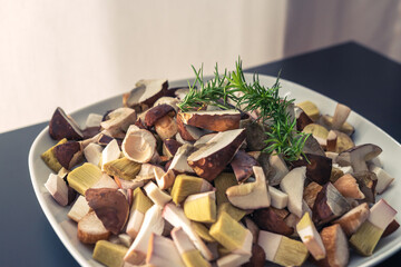 White plate full of chopped, fresh bay boletes and boletus, decorated with green twig, standing on a black table. Bright background.