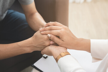 Asian young man, male suffers in mental need to therapy with psychologist while sitting on couch, consult with psychiatrist and encouragement patient by touching, holding to make his feel relaxed.