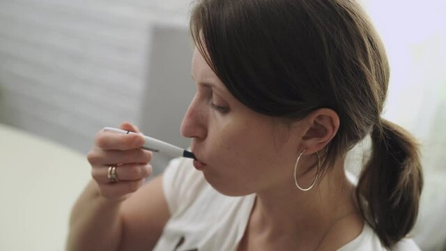 young woman measures her body temperature orally, passes an online medical consultation using a video call on a mobile phone. A sick woman with coronavirus fever, holding a thermometer