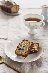 Fototapeta na wymiar Slice of Pound Loaf Cake with raisins on white porcelain plate and cup of tea on greige linen tablecloth. Selective focus