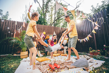Mother and father swing little son holding by hands and legs above plaid with sister at funny birthday celebration in cottage yard in summer