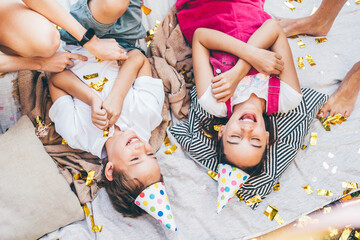 Little children laughing on plaid with confetti and air balloons at birthday celebration in cottage yard in summer. - obrazy, fototapety, plakaty
