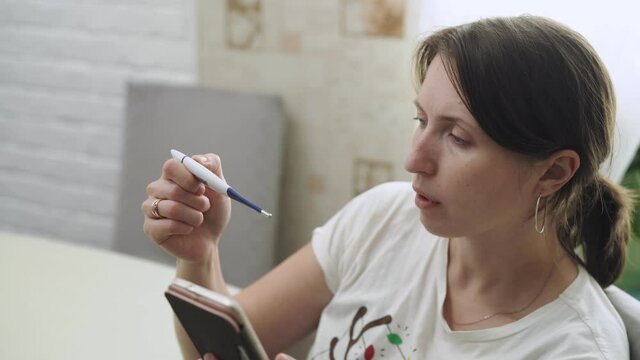 young woman measures her body temperature orally, passes an online medical consultation using a video call on a mobile phone. A sick woman with coronavirus fever, holding a thermometer