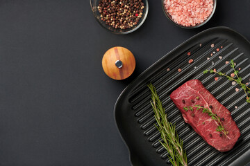 Raw fresh beef top blade steak on grill pan with rosemary and thyme ready for cooking. Marbled meat beef on a frying pan on black background, top view with copy space text.