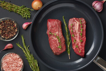 Two raw fresh beef top blade steak on frying pan with rosemary and thyme ready for cooking. Marbled...