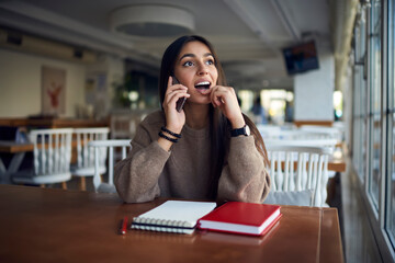 Shocked Middle Eastern female student with education textbook and notepad feeling surprised during cellular conversation in college campus, amazed South Asian woman discussing organization plan
