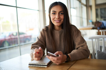 Portrait of cheerful international student with education textbook and modern mobile device smiling at camera during school time for learning and studying, happy Middle Eastern blogger