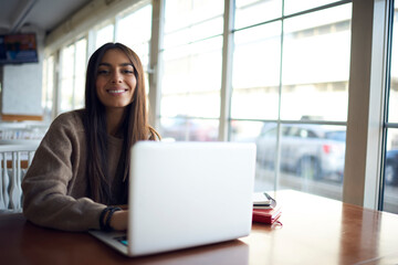 Portrait of cheerful female IT professional with modern laptop computer smiling at camera during distance job in coworking space, happy Caucasian woman with netbook enjoying freelance lifestyle