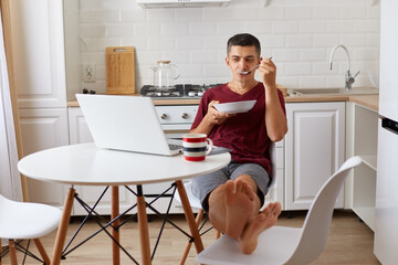 Relaxed handsome man wearing maroon casual t shirt sitting at table, putting feet on chair, eating soup and watching film, enjoying breakfast during weekend or while having break from online work.