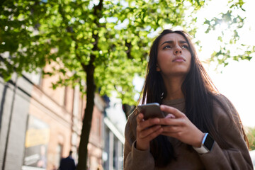 Middle Eastern hipster girl with cellular technology thoughtful looking away and thinking, millennial female travel blogger feeling pondering on text idea for travel website using cellphone device