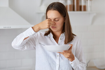 Indoor shot of woman standing with plate in hands, does not want eat soup, feeling bad smell,...