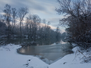Calm snowy winter scene with river, snow covered riverbanks and naked trees