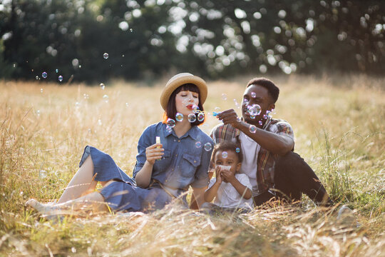 Joyful mixed race family of three sitting together on grass and blowing soap bubbles. Happy parents playing with little cute son on nature. Picnic and fun time.