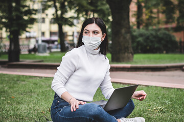 Fototapeta premium A young girl in a protective mask works in the park on a laptop and with a phone