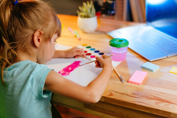 Little girl elementary student drawing studying online from home using laptop sitting at the desk