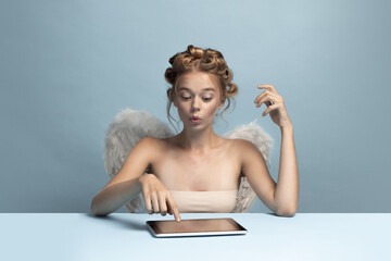 Portrait of young beautiful girl in image of angel with wings using gadget, tablet isolated on blue gray studio background. Concept of beauty, purity, tenderness, inner world