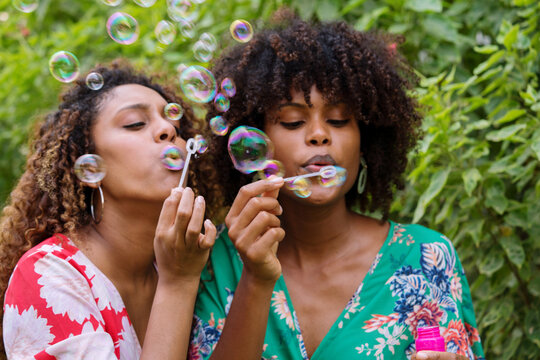 Two Happy Black woman with curly hair having fun blowing soap bubbles outdoors in the garden
