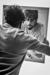 portrait of a young man with beard and curly hair, who looks at himself while holding a mirror in his hands