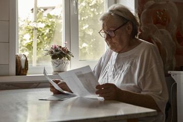 Elderly woman sitting at table in kitchen at home holds domestic bills, feels concerned forgot to...