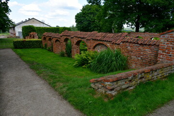 A close up on a wall made out of red brick with some arches and decorative elements covered with vines, shrubs, and grass located in the middle of a well maintained public park seen on a sunny day