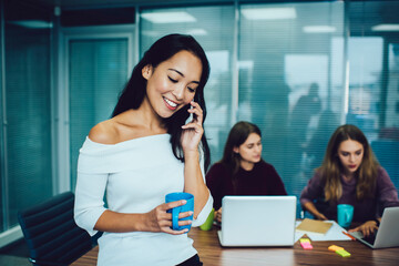 Happy Asian employee with caffeine beverage using cellular device for friendly calling during coffee break in office interior, smiling executive manager discussing brainstorming while phoning