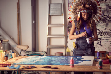 A young female artist with a war bonnet in the studio is focused on a work on a new painting. Art, painting, studio