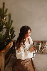 A sweet happy young woman in a cozy sweater is resting looking at a gift box sitting on the sofa by the Christmas tree during the winter holidays at home