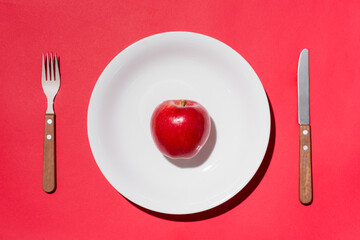 Top view of red apple on white plate with knife and fork on red background