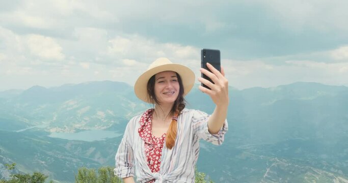 Happy woman traveler having video call using smartphone in mountains. Woman in hat sharing travel adventure with family, friends. Girl taking selfie video, photo with mountains view for social media