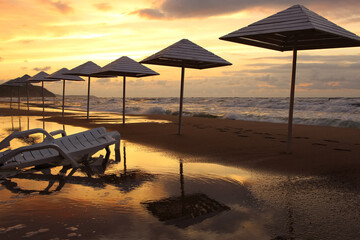 Umbrellas and sun loungers on wet sand by the beach in stormy weather in sunset light and sun reflections 