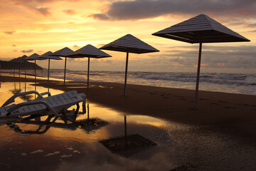 umbrellas and sun lounger on wet sand by the beach in stormy weather in sunset light and sun reflections 