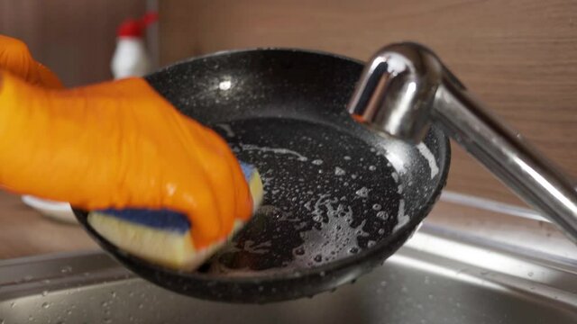 A man's hand in a glove with a sponge washes a dirty frying pan. Wash the pan with a sponge and detergent