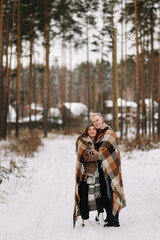 A happy married couple in love is hiding under a plaid blanket, warming up while walking traveling in the winter forest in nature in cold weather, selective focus