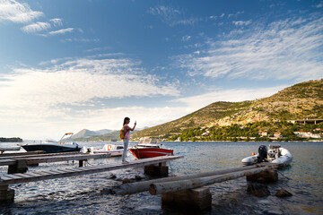 Young tourist woman standing on wooden berth with small boats near and making photo with her mobile of fantastic croatian coast view. Travel Dubrovnik, Croatia.
