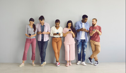 Indoor shot of diverse international group of happy young millennial people leaning on wall, looking at screens of mobile phones, connecting to free wi fi, using messengers, watching online streams