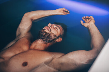 Handsome young bearded man floating in tank filled with dense salt water used in meditation,...